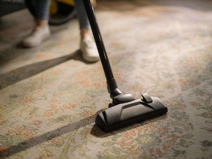A person using a vacuum cleaner to deep clean a patterned area rug with floral designs, in a room with wooden floors and natural lighting. The vacuum cleaner, equipped with a wide cleaning head, is be