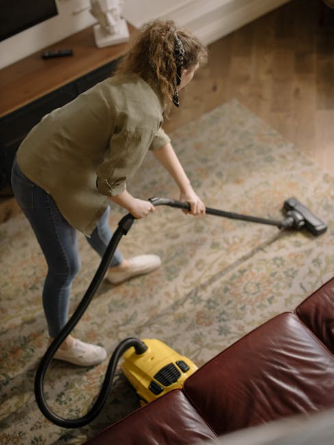 A person using a vacuum cleaner to deep clean a patterned area rug with floral designs, in a room with wooden floors and natural lighting. The vacuum cleaner, equipped with a wide cleaning head, is be