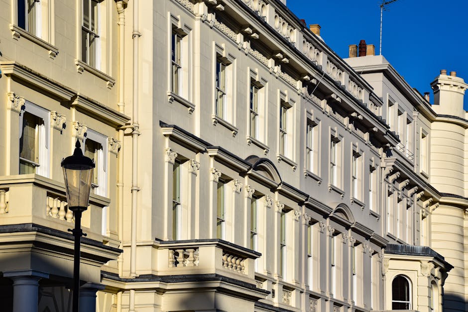 Photograph of a row of elegant, white Victorian-style residential buildings with intricate architectural details, large windows, and balconies. The buildings are set along a paved street lined with le