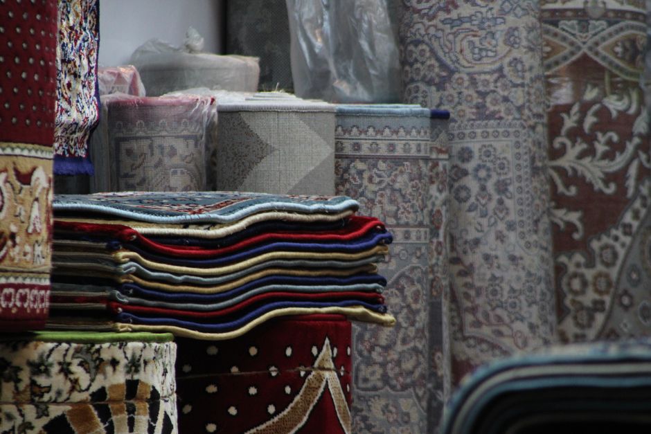 A person standing outdoors on a grassy area, partially visible from the waist down, holding a rolled-up patterned rug with a traditional design featuring shades of orange, cream, and blue. The rug is 